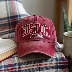 Könnte beinhalten: Eine kastanienbraune Baseballkappe mit Used-Look, die das Wort CUSTOM in großen, weiß umrandeten roten Blockbuchstaben über dem Wort COLLEGE in kleineren weißen Blockbuchstaben zeigt. Die Kappe hat einen gebogenen Schirm und sechs Paneele.