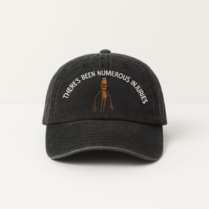 May include: Black baseball cap with the text "THERE'S BEEN NUMEROUS INJURIES" in white, arched above a cartoon character. The character is brown with a light brown face and arms. The cap has a curved brim.
