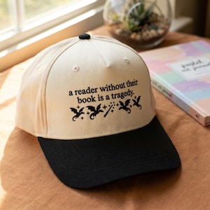 May include: A baseball cap with a cream-colored crown and a black brim. The cap features the text "a reader without their book is a tragedy" with dragon and wand graphics.