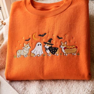 May include: An orange crewneck sweatshirt featuring an embroidered Halloween scene. The design includes four Corgi dogs dressed in costumes: a mummy, a ghost, a witch with a hat and cape, and a hot dog. Bats, a crescent moon, and stars are above.
