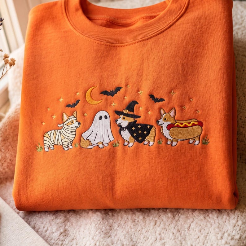May include: An orange crewneck sweatshirt featuring an embroidered Halloween scene. The design includes four Corgi dogs dressed in costumes: a mummy, a ghost, a witch with a hat and cape, and a hot dog. Bats, a crescent moon, and stars are above.