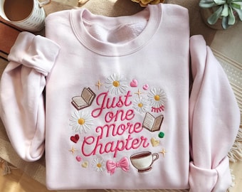 May include: A light pink crewneck sweatshirt featuring embroidered text that reads Just one more Chapter. The design includes white daisies, open books, a coffee cup, a pink bow, and small hearts, all arranged in a circular pattern on the chest.