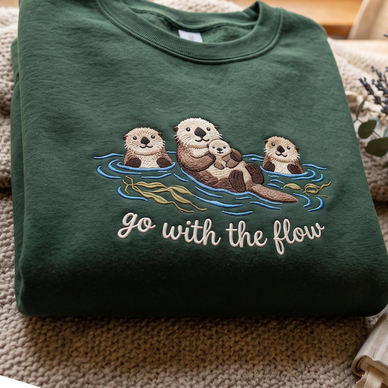 Puede incluir: Una sudadera de cuello redondo verde oscuro con un dise&ntilde;o bordado de tres nutrias marrones nadando en agua azul con algas verdes. La nutria central sostiene a una cr&iacute;a. Debajo del bordado, el texto "go with the flow" aparece en color blanco.
