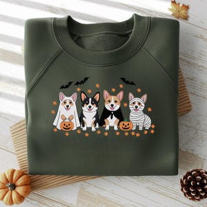 Może przedstawiać: Ciemnozielona bluza z kapturem z motywem Halloween. Projekt przedstawia cztery corgi w kostiumach: ducha, wampira, mumię i jednego z peleryną. Każdy corgi stoi obok dyni. Zawiera również nietoperze i pomarańczowe gwiazdy.