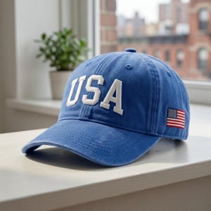 May include: A blue baseball cap with the word "USA" in white block letters across the front. An American flag patch is on the side. The cap is sitting on a white surface.