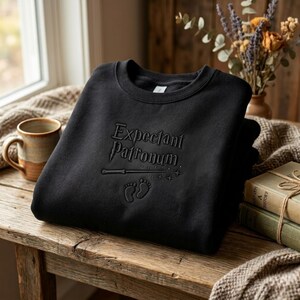 May include: Black sweatshirt with the embossed text "Expectant Patronum", a wand, baby footprints, and stars. The sweatshirt is folded on a wooden surface, with a mug and books in the background.