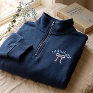 Könnte beinhalten: Marineblaues Sweatshirt mit Viertelreißverschluss und dem Wort "radiology" in hellblauer Schrift mit einer rosa Schleife gestickt. Das Sweatshirt hat einen gerippten Kragen und Bündchen sowie einen silbernen Reißverschluss. Im Hintergrund ein Buch mit dem Titel "Radiology & Imaging".