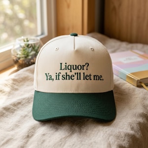 May include: A baseball cap with a cream-colored crown and a dark green brim. The cap features the text "Liquor? Ya, if she'll let me" in dark green lettering. The cap has a dark green button on top.