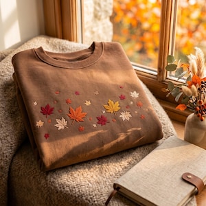 May include: A brown crewneck sweatshirt featuring an embroidered pattern of scattered maple leaves in shades of red, orange, yellow, and cream across the chest. The garment has a ribbed collar and long sleeves, showcasing a detailed leaf design.