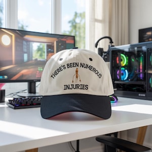 May include: Cream-colored baseball cap with a navy blue brim. The cap features the text "THERE'S BEEN NUMEROUS INJURIES" in black, with a cartoon graphic. The cap is sitting on a white desk.
