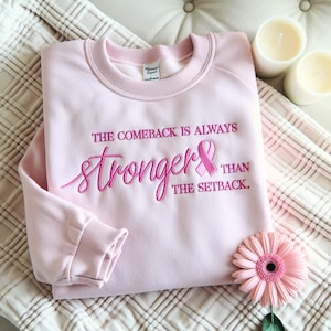 Könnte beinhalten: Hellrosa Sweatshirt mit der gestickten Aufschrift "THE COMEBACK IS ALWAYS STRONGER THAN THE SETBACK" und einem rosa Band. Eine rosa Gerbera und zwei weiße Kerzen sind ebenfalls zu sehen.