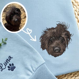May include: Light blue sweatshirt featuring a detailed embroidered portrait of a black dog. A smaller circular image of the dog is in the upper left corner. The name "Luna" and a paw print are embroidered on the lower left.