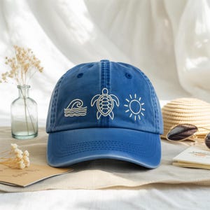 Pode incluir: Boné de beisebol azul com aba curva. O boné apresenta desenhos bordados em branco de ondas, uma tartaruga e um sol. O chapéu é feito de um tecido macio, adequado para uso casual.