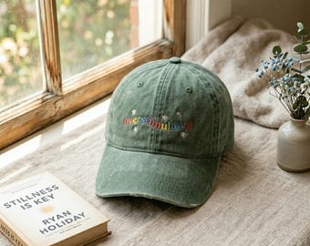 May include: A sage green baseball cap with a distressed finish, featuring the embroidered word "overstimulated" in a rainbow gradient font, accented by small white star icons stitched around the text on the front panel.
