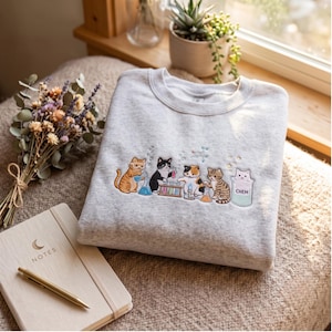 Pode incluir: Uma camisola de gola redonda cinzento-claro com um design bordado de cinco gatos a realizar experiências científicas com gobelés, tubos de ensaio e um bico de Bunsen. Um gato está dentro de um gobelé com a etiqueta CHEM.