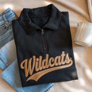 Könnte beinhalten: Ein schwarzes Sweatshirt mit einem Kragen mit Reißverschluss und dem Wort "Wildcats" in goldener Schrift gestickt. Das Sweatshirt ist gefaltet, mit einer Jeans und einer weißen Tasse im Hintergrund. Das Sweatshirt ist aus weichem Material.