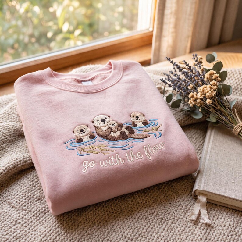 Puede incluir: Una sudadera rosa claro con un dise&ntilde;o bordado de tres nutrias marinas nadando en agua azul. La nutria central sostiene a una cr&iacute;a, mientras que el texto "go with the flow" est&aacute; cosido en letras cursivas blancas debajo de las nutrias.