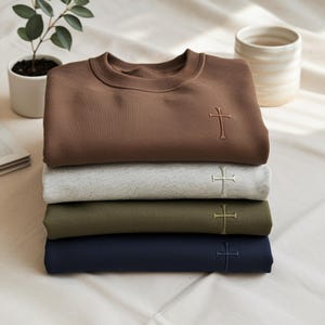 Könnte beinhalten: Ein Stapel aus vier Rundhals-Sweatshirts in Braun, meliertem Grau, Olivgrün und Marineblau. Jedes Sweatshirt ist mit einem kleinen, gestickten Kreuzmotiv auf der rechten Brust versehen, das bei allen vier gefalteten Kleidungsstücken einheitlich platziert ist.