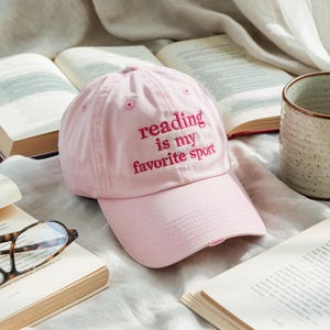 Op de afbeelding: Lichtroze baseballpet met de geborduurde tekst "reading is my favorite sport" in donkerder roze. De pet is omgeven door open boeken en een keramische mok.