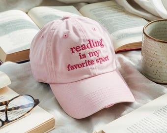 May include: A light pink baseball cap with the embroidered text "reading is my favorite sport" in a darker pink. The cap is surrounded by open books and a ceramic mug.