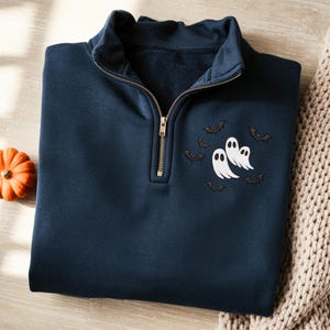 Op de afbeelding: Marineblauwe sweatshirt met een kwartrits en een gouden rits. De sweatshirt heeft een geborduurd ontwerp van drie witte geesten en verschillende zwarte vleermuizen, perfect voor Halloween. Een kleine oranje pompoen is zichtbaar op de achtergrond.