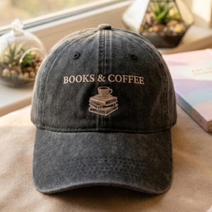 Może przedstawiać: Ciemnoszara czapka bejsbolówka z haftowanym napisem "BOOKS & COFFEE" nad stosikiem książek i filiżanką kawy. Czapka wykonana jest z zużytej, wyblakłej tkaniny i ma zakrzywiony daszek.
