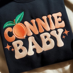 May include: Black sweatshirt with embroidered text "CONNIE BABY" in orange and peach colours, featuring a peach graphic with green leaves. The design includes orange star accents.