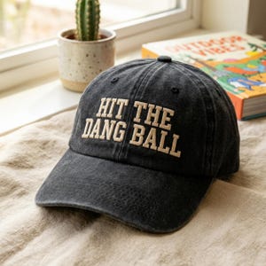 Può includere: Un cappellino da baseball nero con la scritta ricamata "HIT THE DANG BALL" in crema. Il cappellino è realizzato in tessuto usurato e ha una visiera curva. Un piccolo cactus in vaso è visibile sullo sfondo.