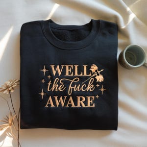 Pode incluir: Camisola preta com bordado dourado "WELL the fuck AWARE", adornada com estrelas e motivos florais. Uma caneca de cerâmica e flores secas estão no fundo.