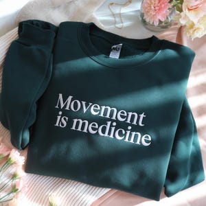 May include: A dark green sweatshirt with the phrase "Movement is medicine" in white lettering. The sweatshirt is folded on a light pink and white striped surface, with flowers and a necklace in the background.
