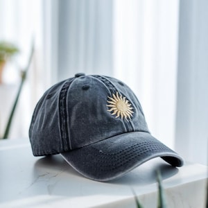 May include: A dark grey baseball cap with a curved peak. The cap has a gold embroidered sun motif on the front. The hat is made of a soft, worn material.