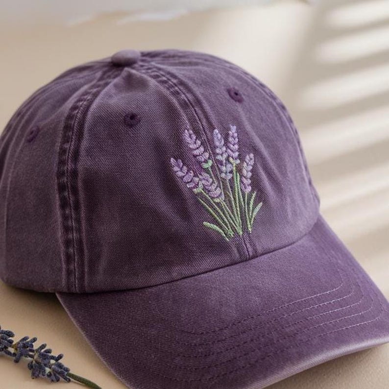 Op de afbeelding: Een paarse baseballcap met een gebogen klep. De cap heeft een geborduurd lavendelontwerp in paarse en groene tinten op de voorkant. De cap is gemaakt van een zachte, getextureerde stof.