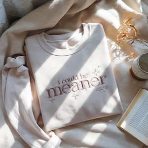 May include: A beige sweatshirt with the embroidered phrase "i could be meaner" in a rose-colored thread. The sweatshirt has a crew neck and long sleeves. The text is surrounded by small starburst designs.