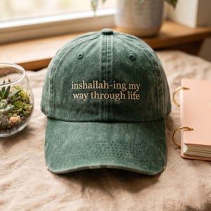 May include: A distressed green baseball cap with the embroidered phrase "inshallah-ing my way through life" in white. The cap is made of a soft, worn fabric and features a curved brim. The hat is a casual accessory.
