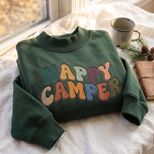 May include: A dark forest green long-sleeve sweatshirt featuring a mock neck collar and ribbed cuffs. The front displays the text "HAPPY CAMPER" in large, puffy, multi-coloured letters with a wavy, retro-inspired font in shades of teal, orange, and pink.