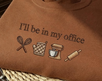 May include: A brown sweatshirt with the embroidered phrase "I'll be in my office" and baking-themed illustrations. The design includes a whisk, oven mitt, stand mixer, and rolling pin.