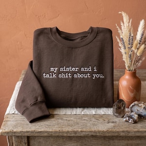 May include: A dark brown crewneck sweatshirt featuring white embroidered text on the chest that reads, my sister and i talk shit about you. The garment has long sleeves with ribbed cuffs and a ribbed neckline, constructed from a solid fabric.