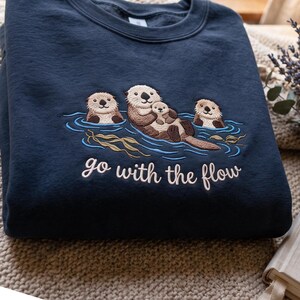 Puede incluir: Una sudadera azul marino de cuello redondo con un bordado de tres nutrias marrones flotando en agua azul con algas verdes. El texto "go with the flow" est&aacute; bordado en letras cursivas blancas debajo de la ilustraci&oacute;n de las nutrias.