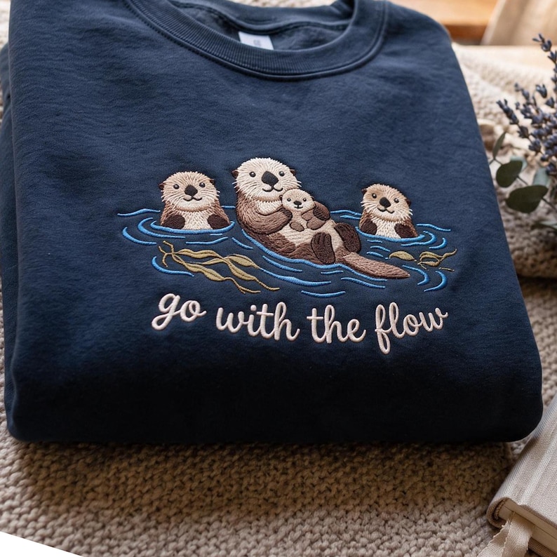 Puede incluir: Una sudadera azul marino de cuello redondo con un bordado de tres nutrias marrones flotando en agua azul con algas verdes. El texto "go with the flow" est&aacute; bordado en letras cursivas blancas debajo de la ilustraci&oacute;n de las nutrias.