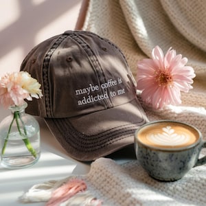 May include: A brown baseball cap with the embroidered text "maybe coffee is addicted to me" sits next to a cup of coffee with latte art. A small vase with flowers and a pink flower are also in the image.