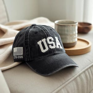 May include: A dark grey baseball cap with the word "USA" in white lettering. The cap has a small American flag and the text "1776-2026" on the side. The hat is on a beige surface.