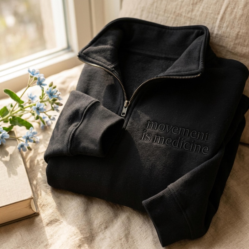 Puede incluir: Sudadera negra con cremallera a media altura y la frase bordada "movement is medicine". La sudadera tiene pu&ntilde;os acanalados y cuello alto. La imagen est&aacute; tomada con luz natural, con un libro y flores en el fondo.