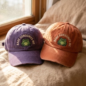 May include: Two baseball caps, one purple and one rust orange, each featuring an embroidered green frog graphic centered above the text RAGE CONSUMES ME. The caps have a structured crown, a curved brim, and a distressed fabric texture throughout.