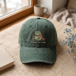 May include: A dark green baseball cap featuring a graphic of a seated frog wearing a red cape and holding a sword. Below the frog, the text reads "I'M BURNT OUT M'LORD" in a simple, light-colored font. The cap has a structured crown and a curved brim.