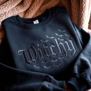 Könnte beinhalten: Ein schwarzes Sweatshirt mit Rundhalsausschnitt, auf dessen Brust das Wort Witchy in einer dunkelgrauen gotischen Schrift gestickt ist, umgeben von einem Muster aus kleinen gestickten Fledermäusen, die über und unter dem Text fliegen.