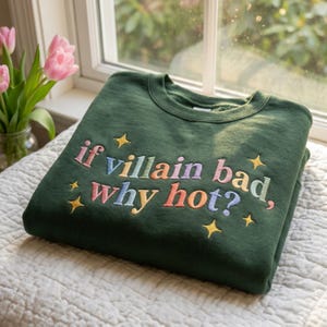 Könnte beinhalten: Dunkelgrünes Sweatshirt mit dem gestickten Satz "if villain bad, why hot?" in bunten Buchstaben und Sternen. Das Sweatshirt liegt gefaltet auf einer weißen Oberfläche in der Nähe eines Fensters mit rosa Tulpen.