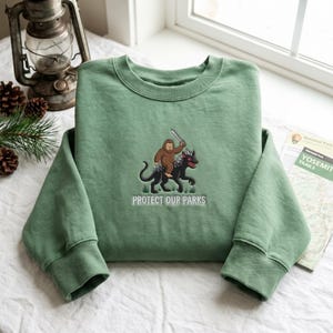 Może przedstawiać: Szałwiowo-zielona bluza z okrągłym dekoltem z haftowaną grafiką przedstawiającą brązowego stwora Bigfoot jadącego na czarnej bestii z rogami i trzymającego miecz, z napisem "PROTECT OUR PARKS" wydrukowanym białymi literami pod ilustracją.