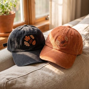 Op de afbeelding: Twee baseballcaps, één in antracietgrijs en één in roestoranje, elk voorzien van een geborduurde cirkelvormige groep herfstbladeren in bruin-, oranje- en crèmetinten. Beide petten hebben een gestructureerde kroon, een gebogen klep en oogjes.
