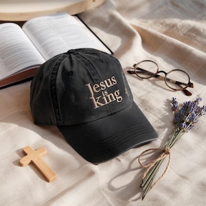 Könnte beinhalten: Eine schwarze Baseballkappe mit dem Text "Jesus is King" in Hellbraun. Die Kappe liegt neben einem Holzkreuz, einer Brille, einem Lavendelstrauß und einem offenen Buch.