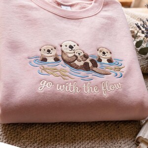 Puede incluir: Una sudadera rosa claro con un dise&ntilde;o bordado de tres nutrias marinas marrones flotando en agua azul con algas color canela. La nutria central sostiene a una cr&iacute;a. Debajo del bordado, el texto dice go with the flow en letra cursiva blanca.